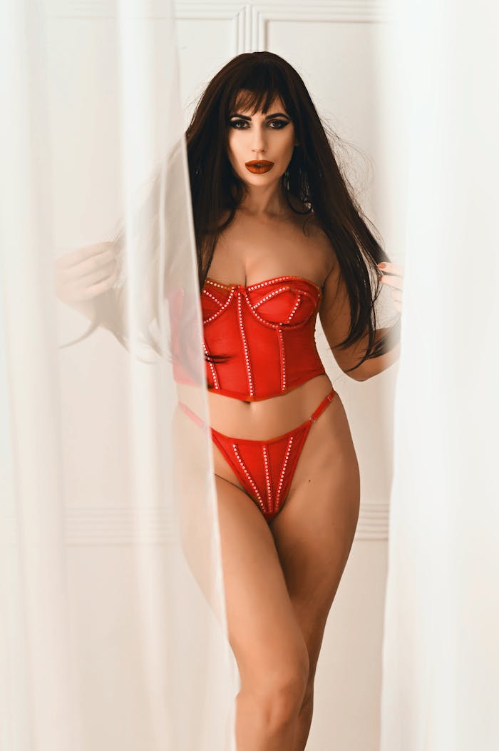 Elegant woman in red lingerie posing behind sheer curtains with a confident look.